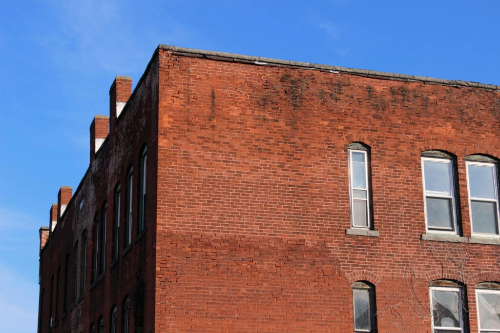 Boarded up windows on abandoned industrial urban brick factory building A corner view of a red brick building showing the side facade with several windows. The structure has a slightly weathered appearance, and the blue sky is visible above with a few scattered clouds.